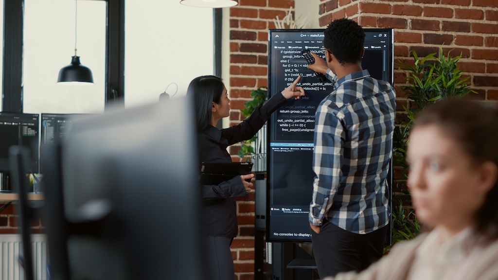 System developers analyzing code on wall screen tv looking for errors comparing results on digital tablet